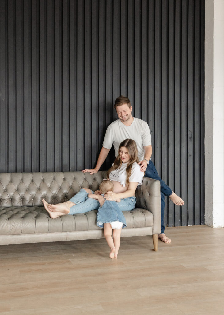 Mom and dad snuggling on the couch while toddler runs toward them, the lumen room denton texas